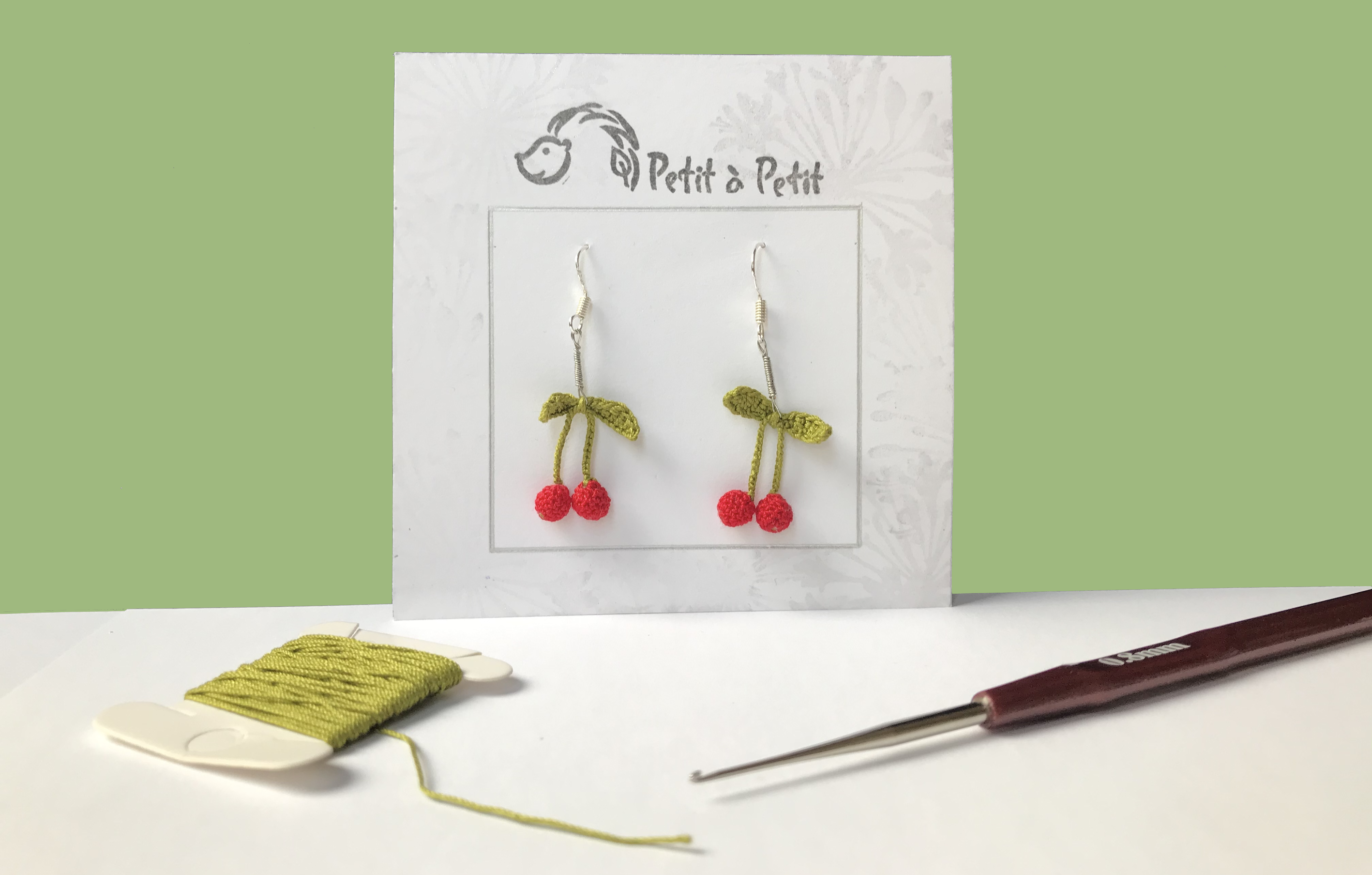 a pair of dangling cherry crochet earings against a matcha green background with a reel of string and crochet hook in the foreground